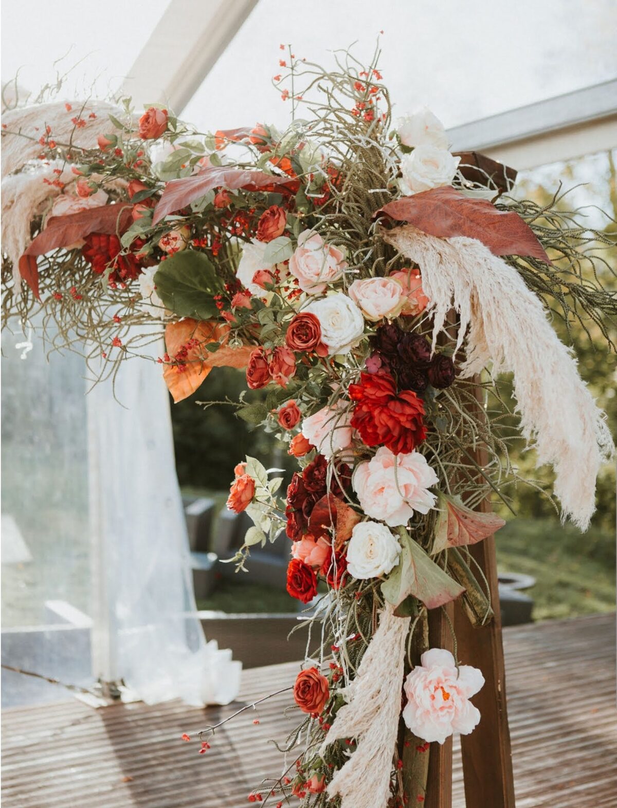 Wedding ceremony flower arch with pampas grass, burgundy and cream-colored flowers in rustic-boho style on a wooden platform surrounded by nature