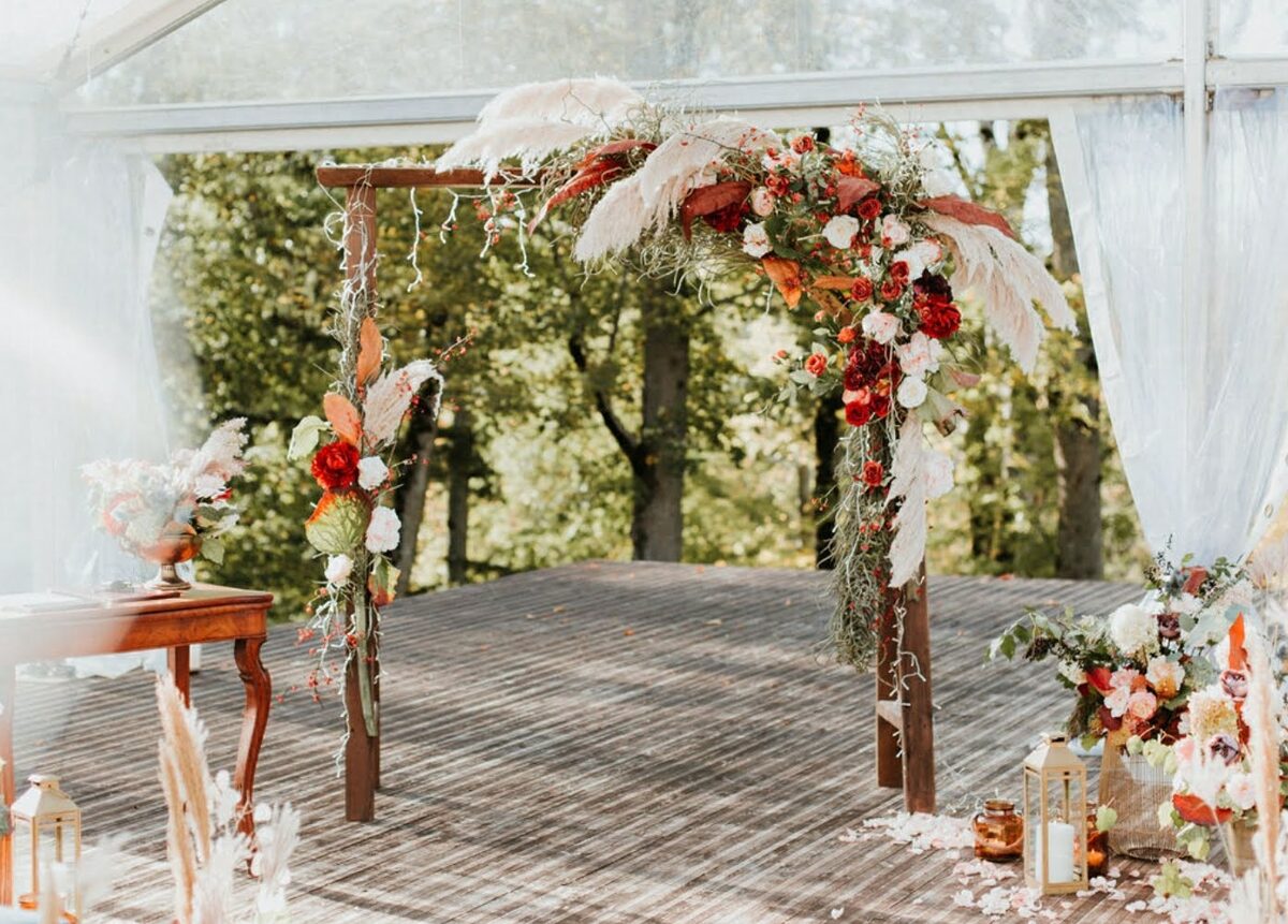 Wedding ceremony flower arch with pampas grass, burgundy and cream-colored flowers in rustic-boho style on a wooden platform surrounded by nature
